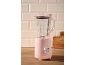 Roze blender sa staklenom posudom i bazom, na drvenoj radnoj ploči sa svetlim pločicama.