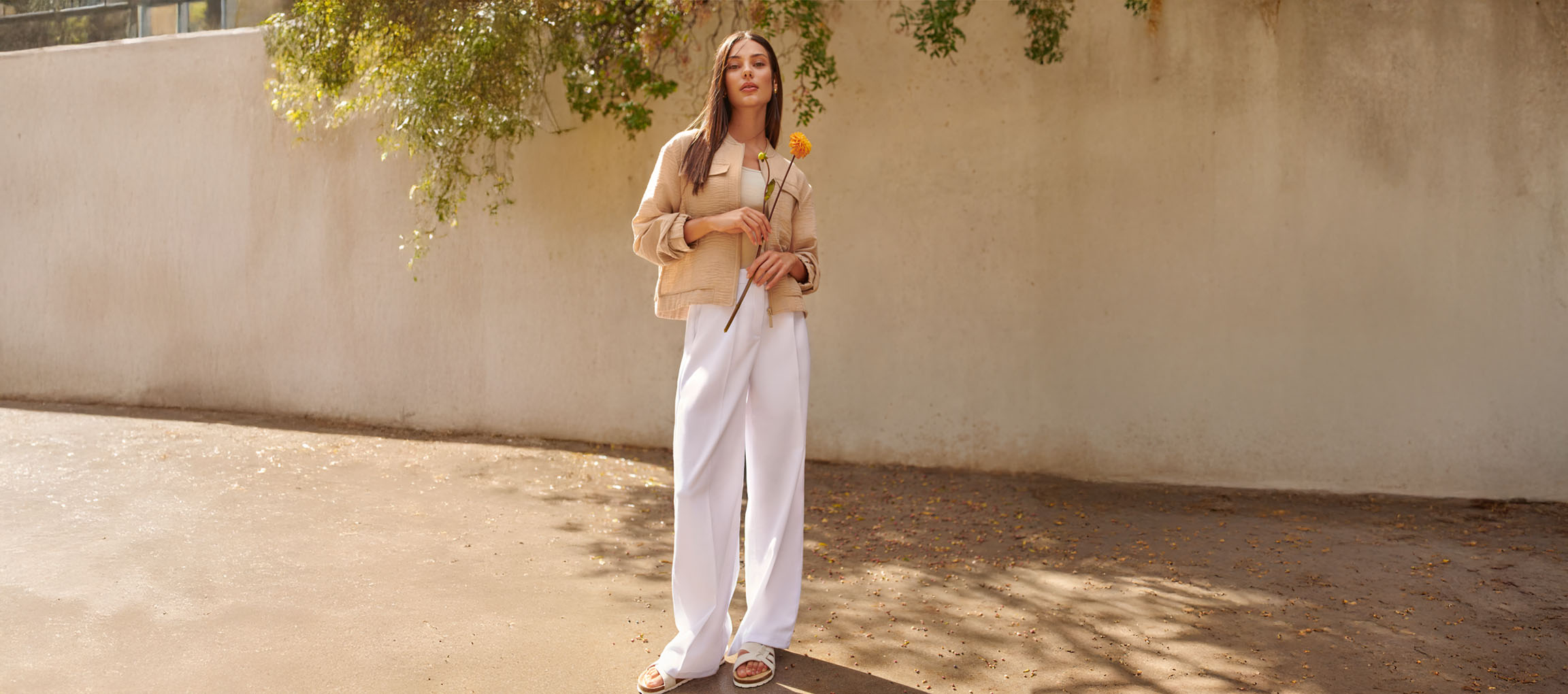 Žena u bež jakni, belim pantalonama i sandalama, drži žuti cvet.