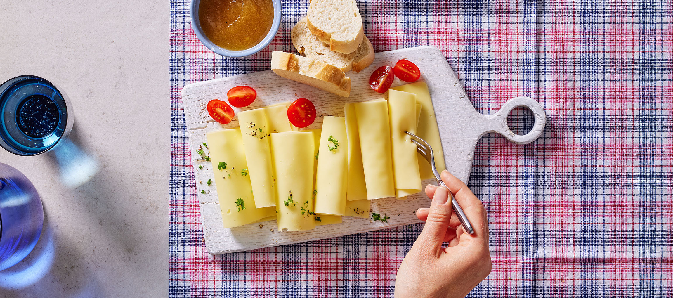 Kriške sira sa čeri paradajzom, hlebom i džemom na kariranom stolnjaku.