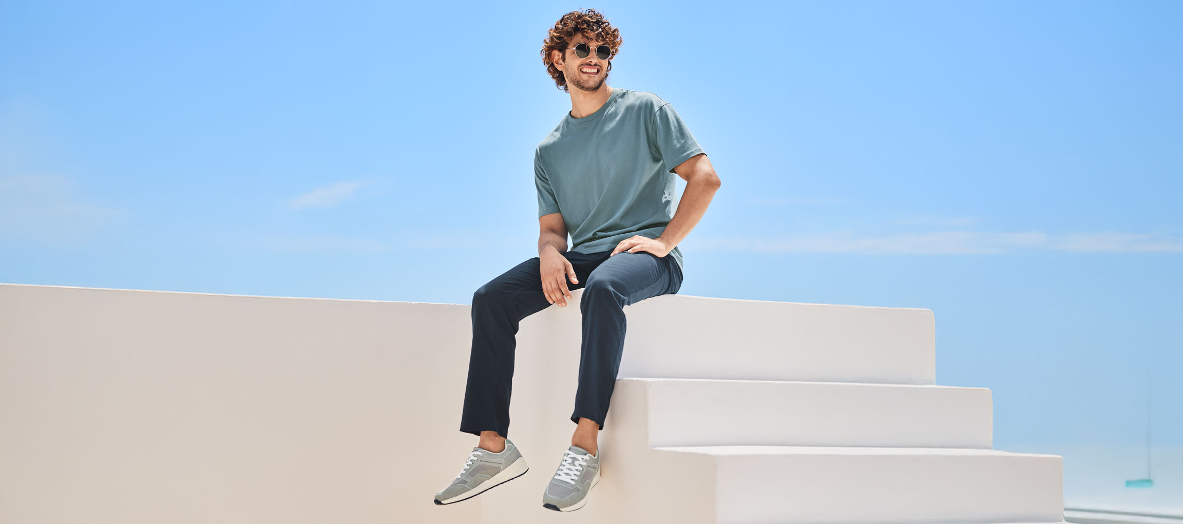 Мушкарац у мајици, панталонама и патикама New Balance седи на белим степеницама.