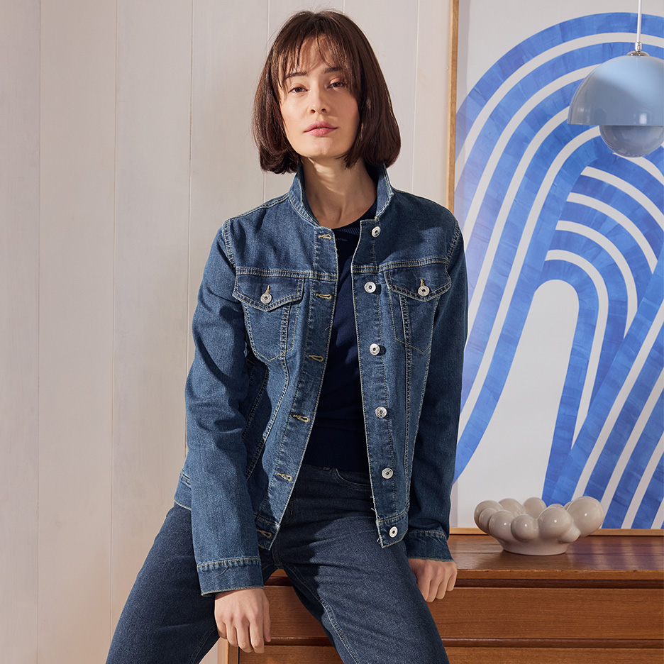 Žena u teksas jakni i farmerkama sedi na drvenoj komodi.