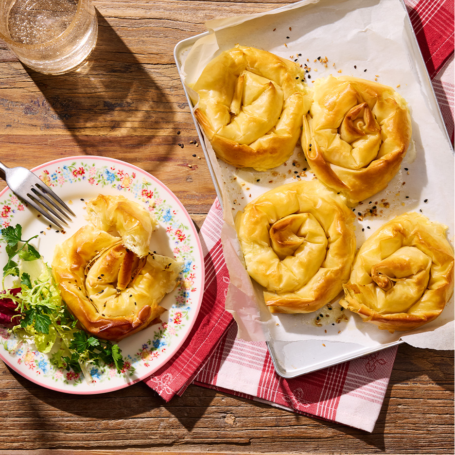 Zlatno-braon spiralne pite sa sirom i susamom, jedna na tanjiru sa salatom, ostale na plehu.