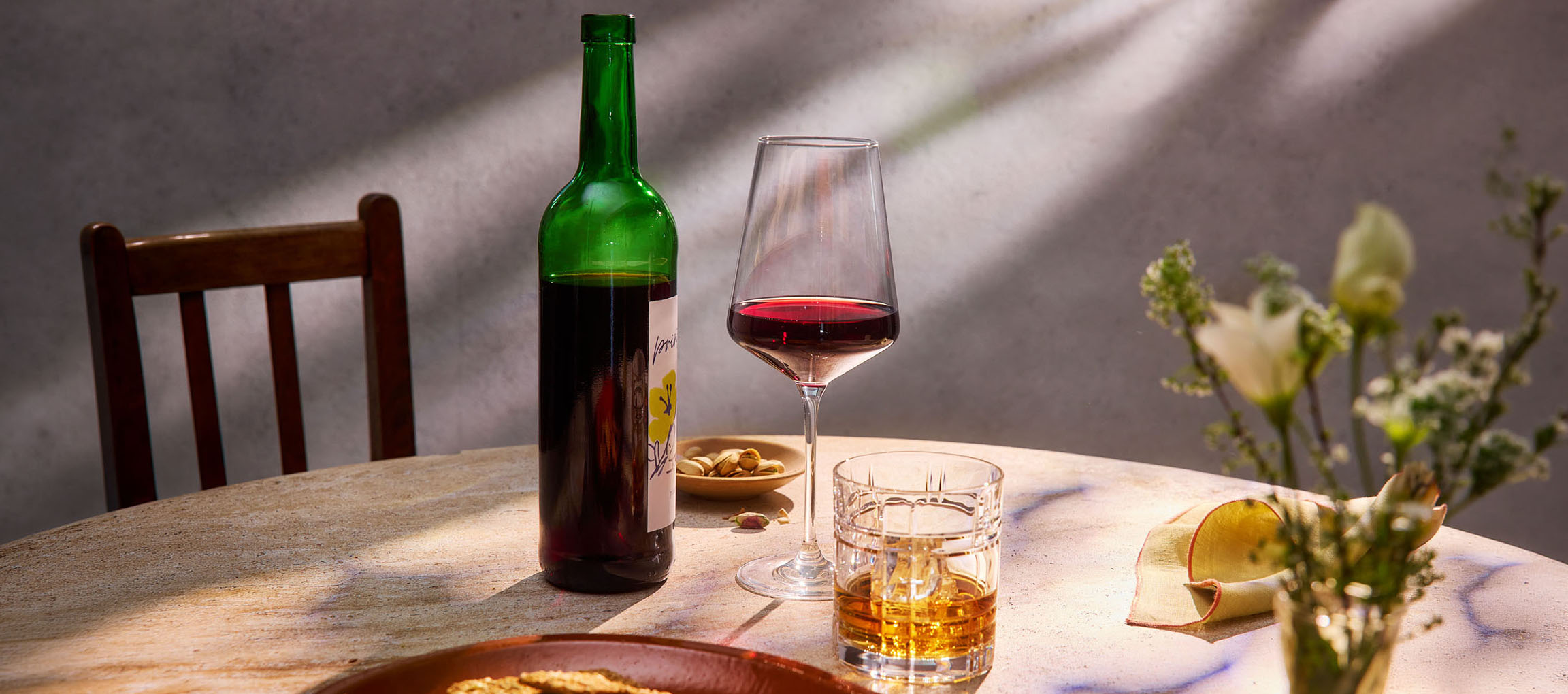 Boca crvenog vina, čaša vina i viski na stolu, sa stolicom i cvećem u pozadini.