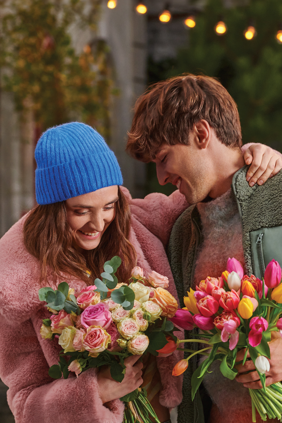 Srećan par sa buketima ruža i lala, u zimskoj odeći, sa svetlima u pozadini.