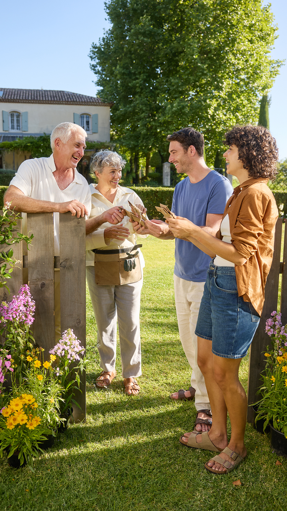 Četvoro ljudi razmenjuje paketiće semena pored drvene ograde i cveća u bašti.