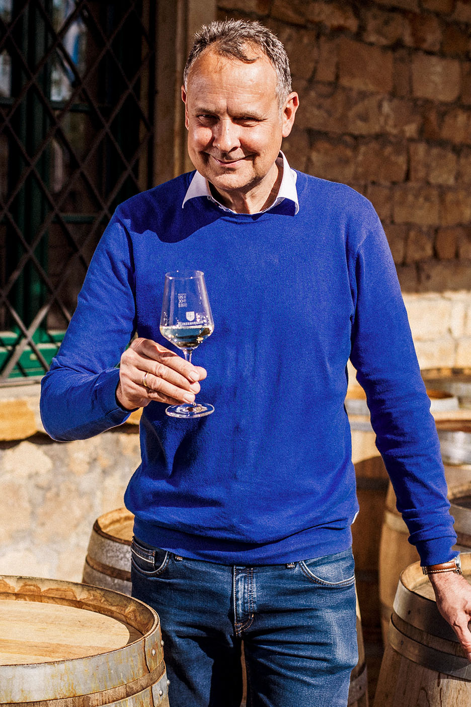 Nasmejani muškarac u plavom džemperu i farmerkama, drži čašu belog vina među drvenim buradima.