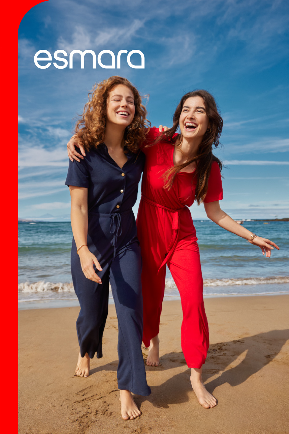 Dve žene u kombinezonima, plavom i crvenom, smeju se na plaži pored mora.