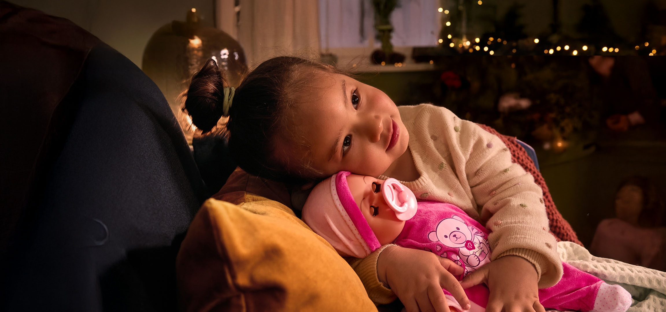 Devojčica sa lutkom i cuclom, udobno smeštena na jastuku.