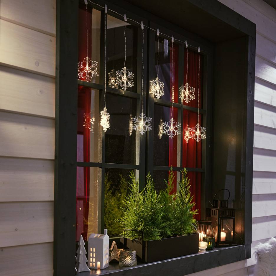 Svetleće pahuljice vise u prozoru sa malim jelkama i svećama na prozorskoj dasci.