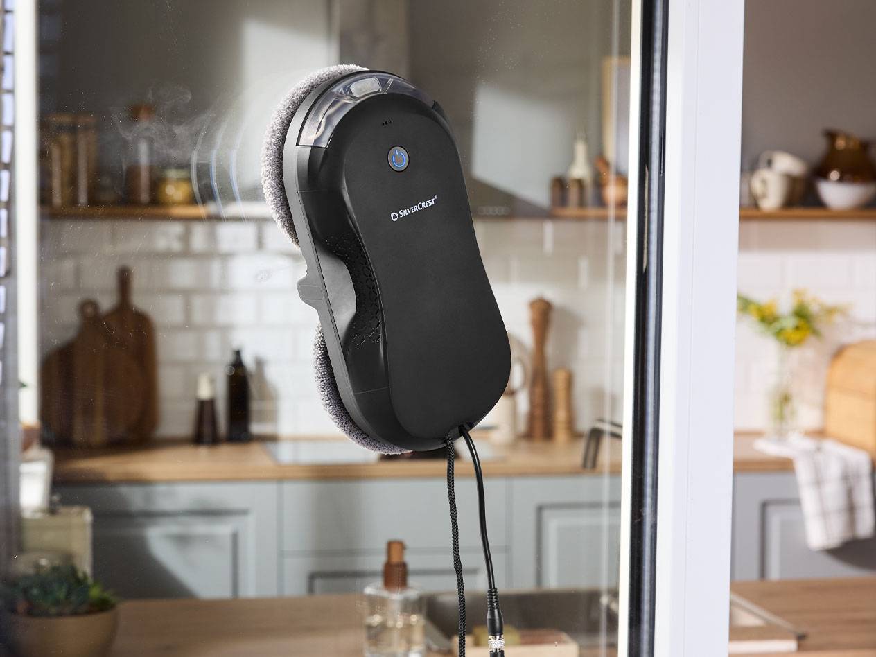 Silvercrest robot za čišćenje prozora na prozorskom staklu sa kuhinjom u pozadini.