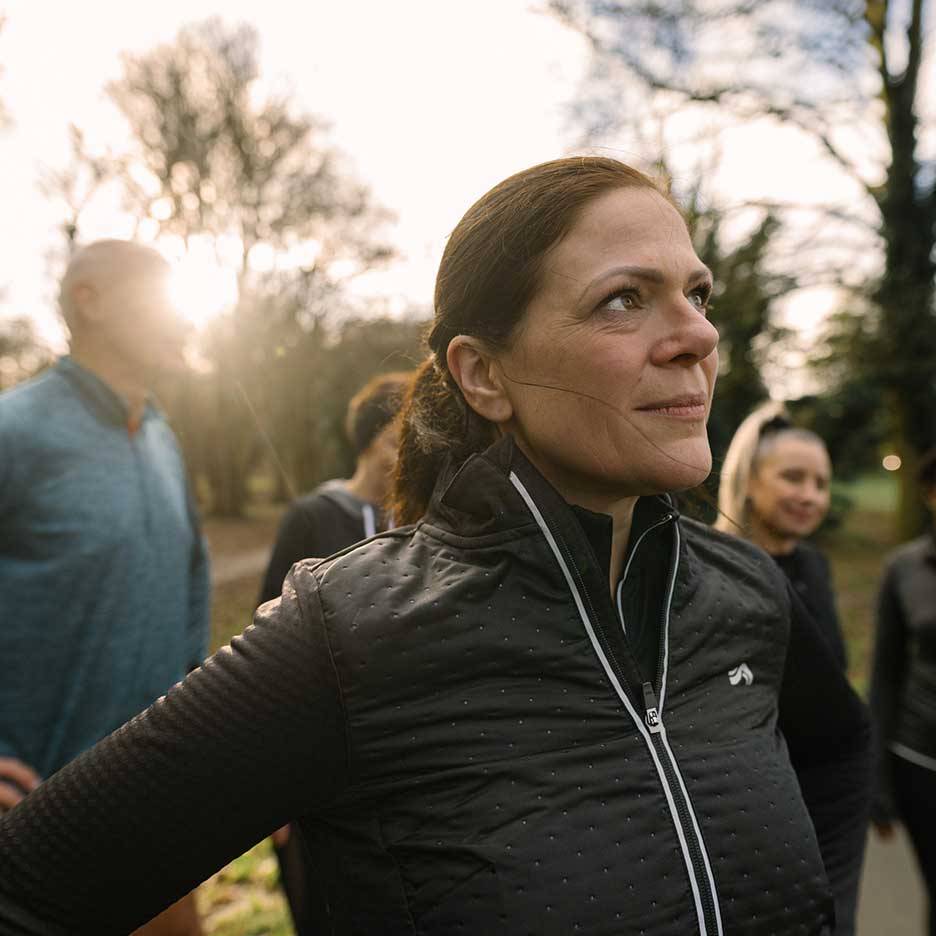 Žena u crnoj sportskoj jakni gleda gore, sa grupom ljudi u pozadini.