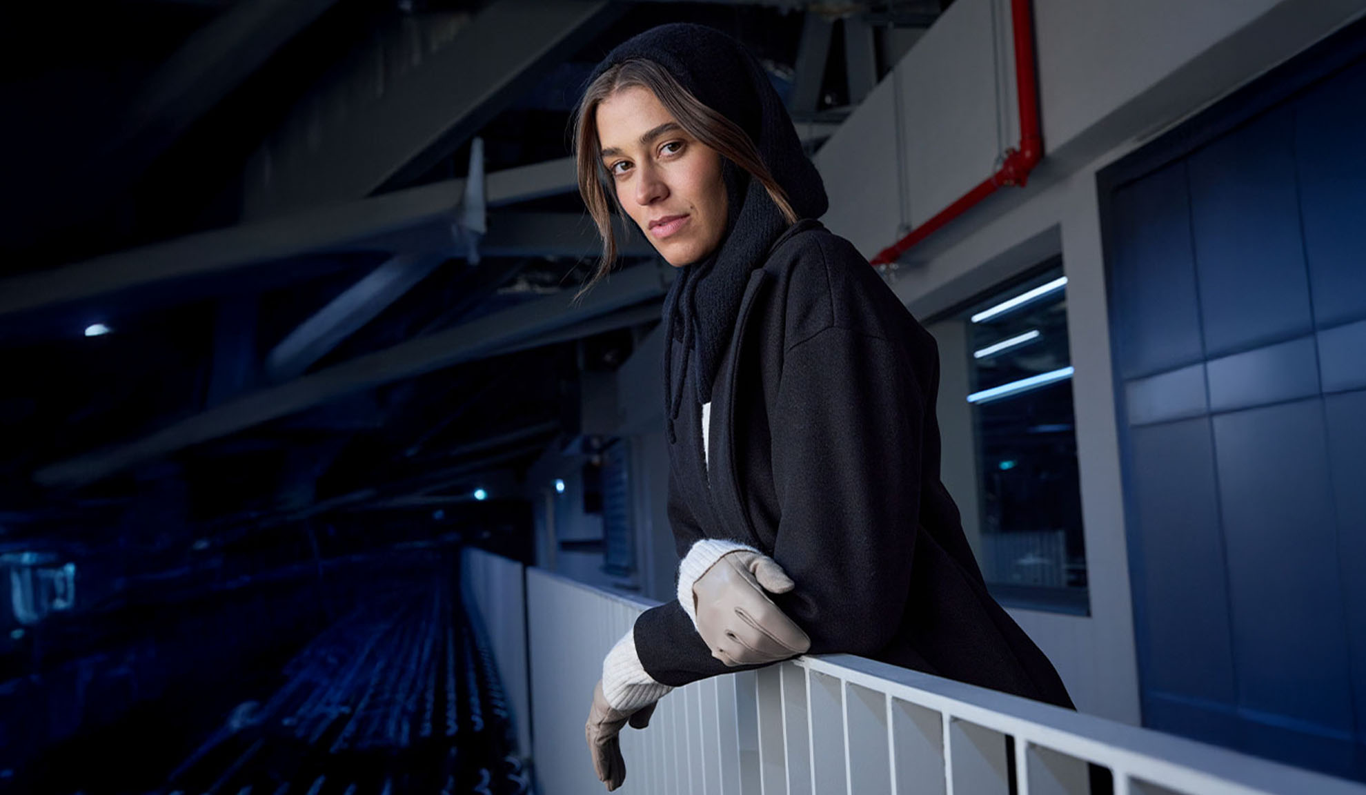 Žena u crnom kaputu i marami sa kapuljačom, naslonjena na ogradu u modernom okruženju.