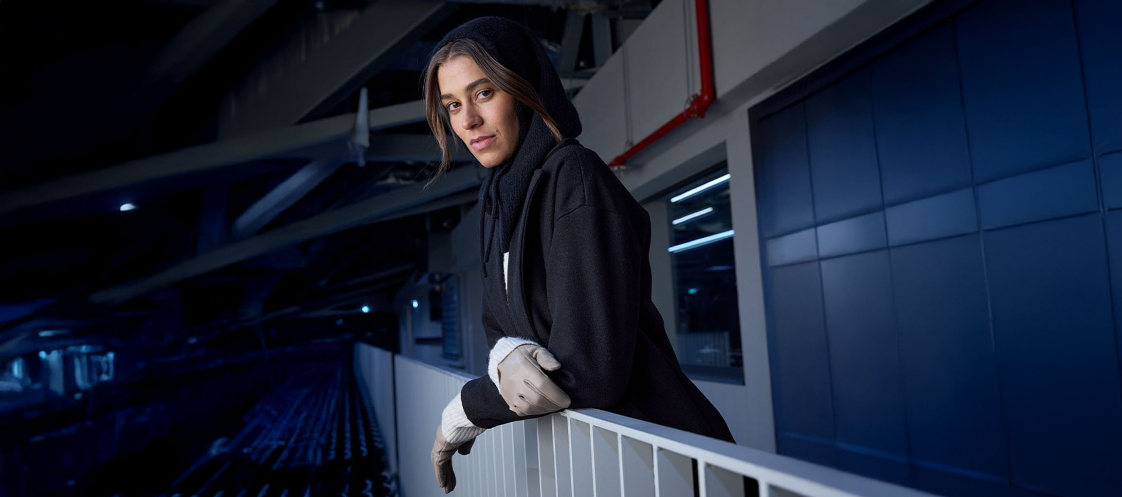 Žena u crnom kaputu i rukavicama naslonjena na ogradu u urbanom okruženju.