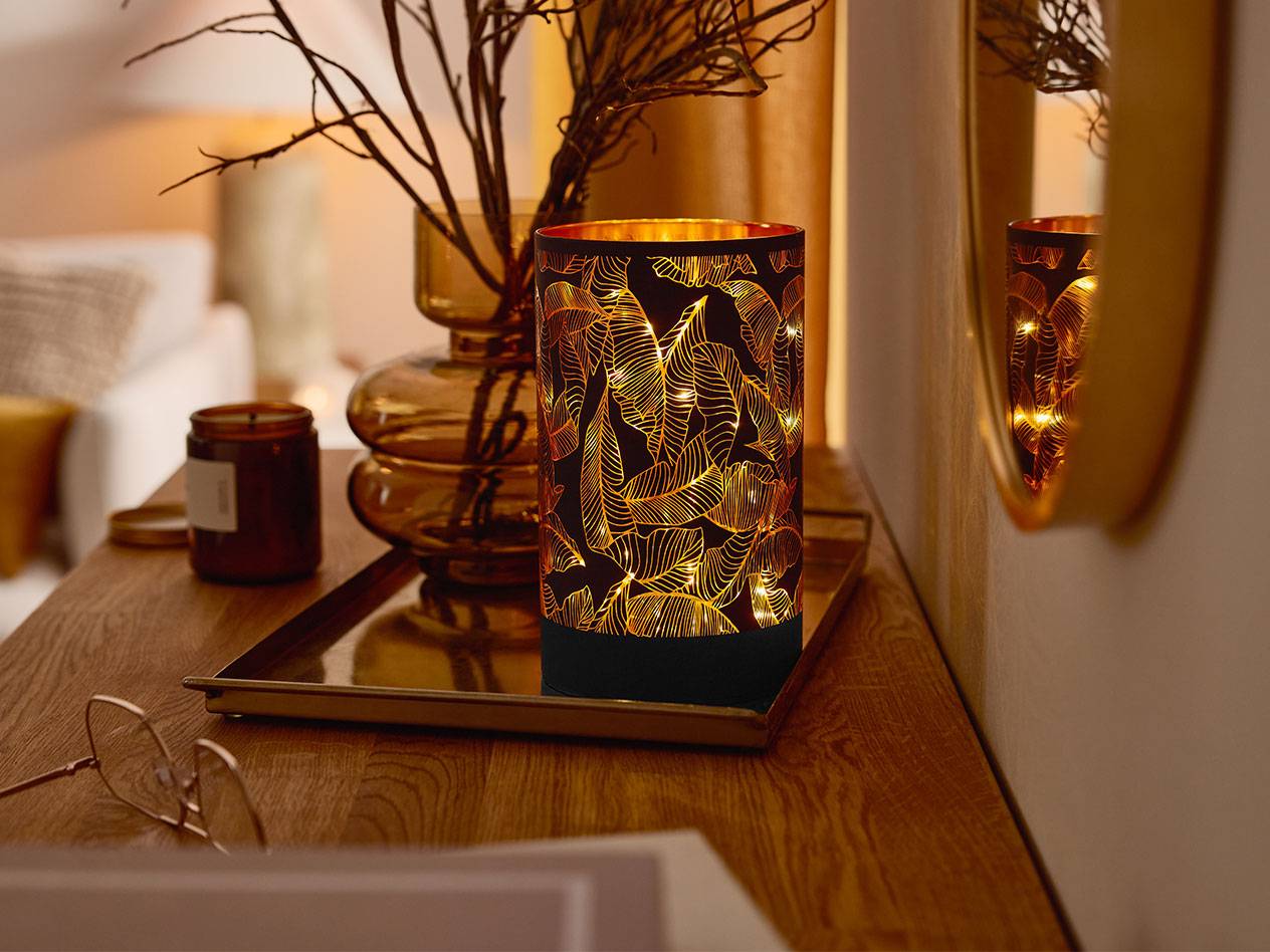 Dekorativna LED lampa sa šarom lišća, vaza sa granama i sveća na drvenom stolu.