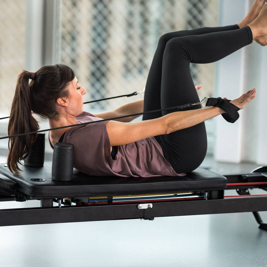 Žena vežba pilates na reformer spravi, u crnoj sportskoj odeći.