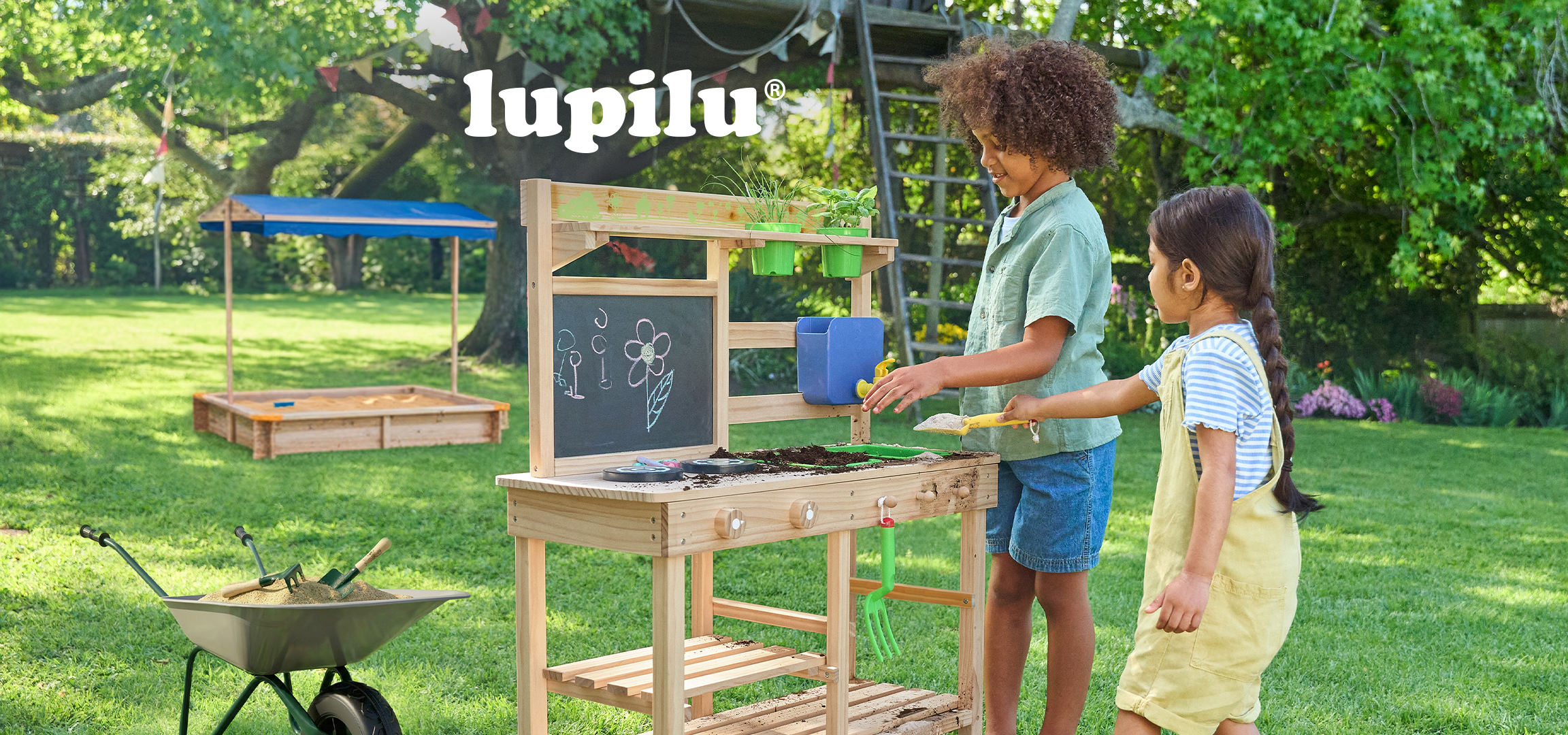 Deca se igraju u bašti sa drvenom kuhinjom za blato i ljuljaškom, u pozadini je peščanik.