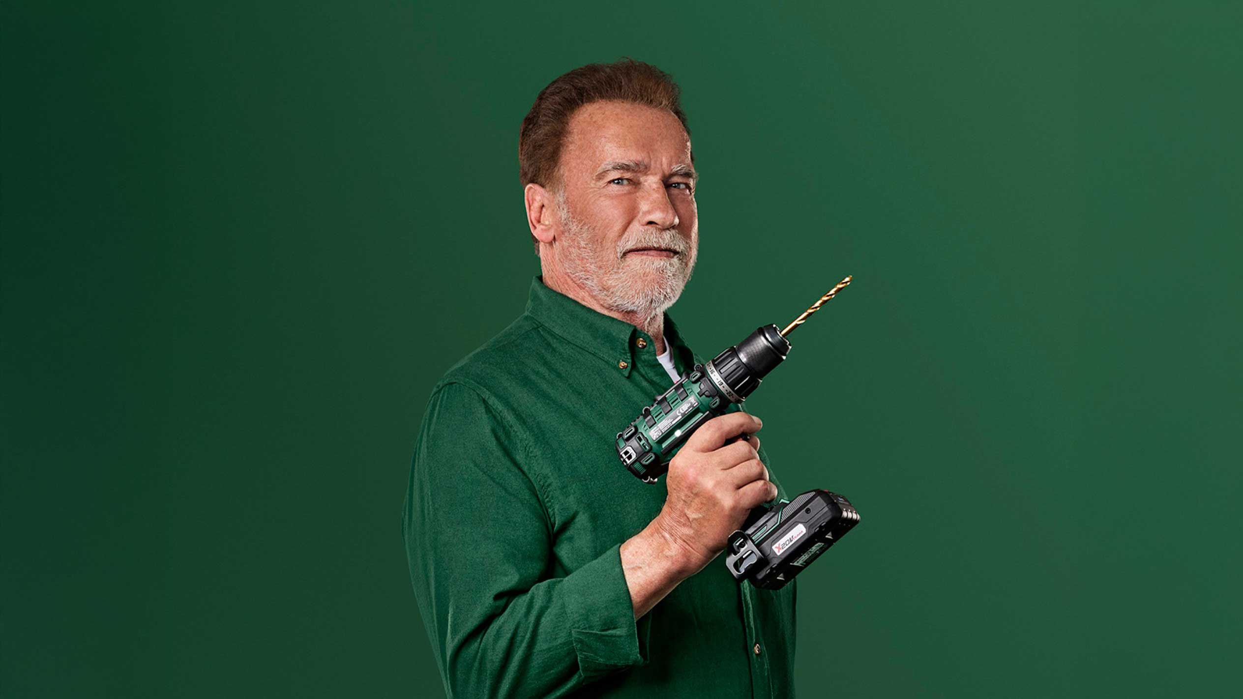Arnold Švarceneger drži zelenu bežičnu bušilicu sa zlatnim burgijom.