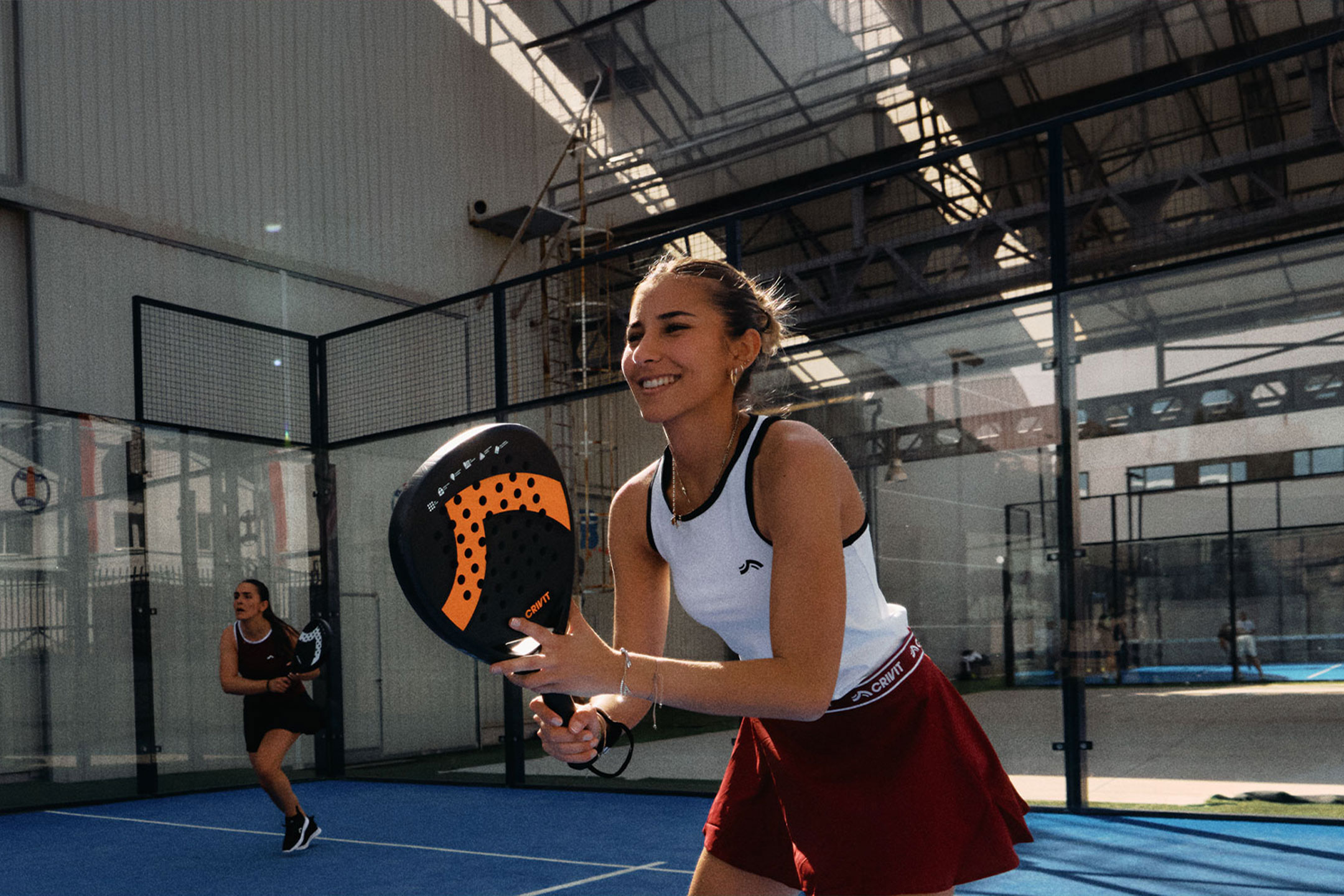 aiLabeling.aiLabelForImageAltText: Dve žene igraju padel tenis, jedna se smeši i drži reket, na plavom terenu.