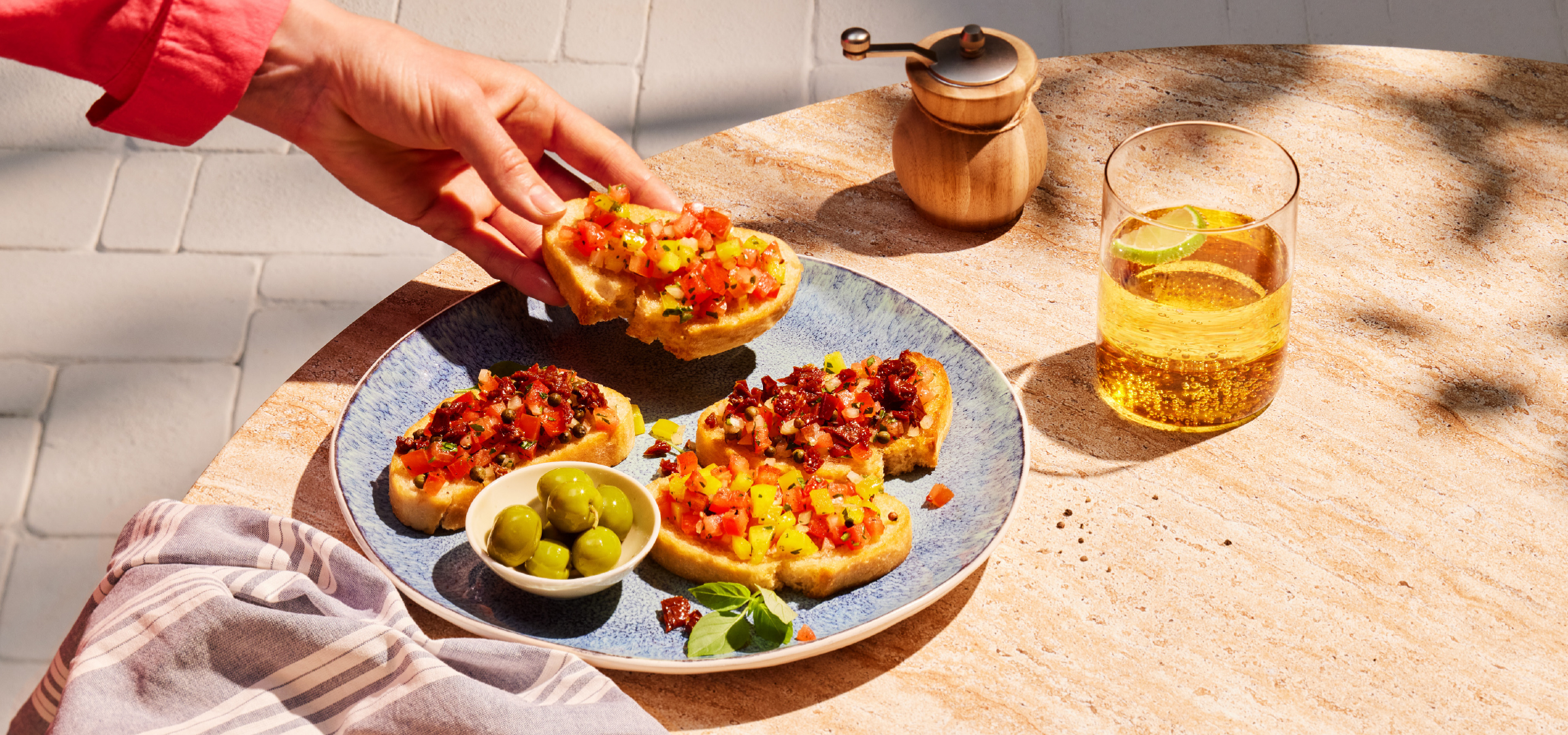 Ruka uzima bruskete sa tanjira, pored su masline, mlin za biber i piće.
