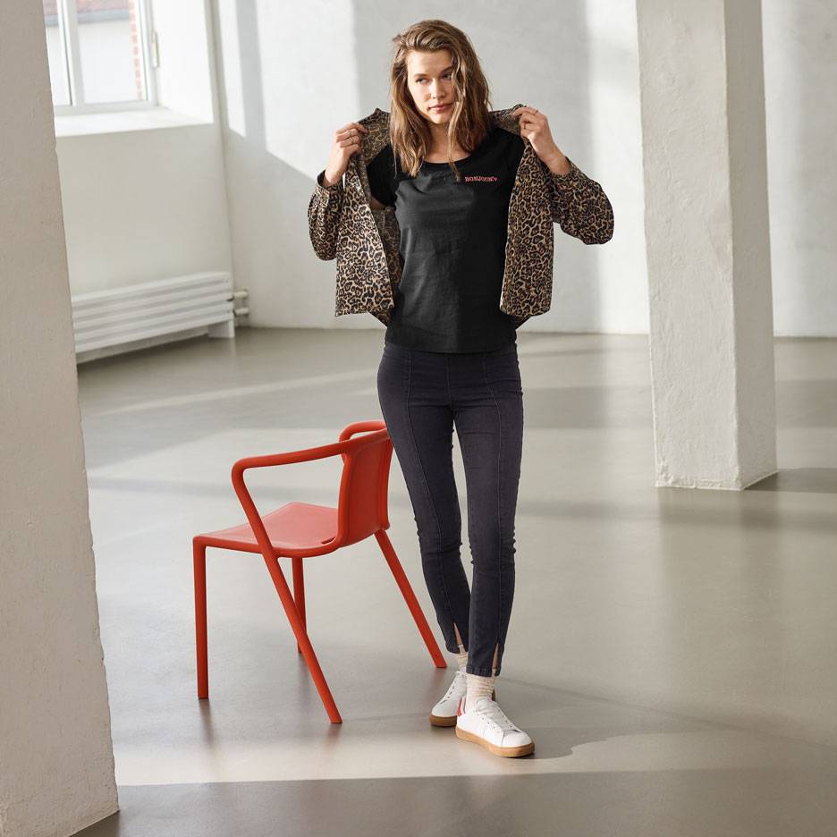 Žena u crnoj majici sa natpisom, farmerkama i jakni sa leopard printom, stoji pored crvene stolice.