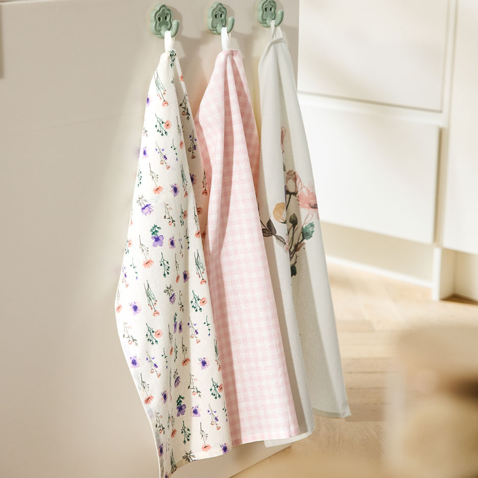 Tri šarene kuhinjske krpe vise na kukama na svetlom zidu.