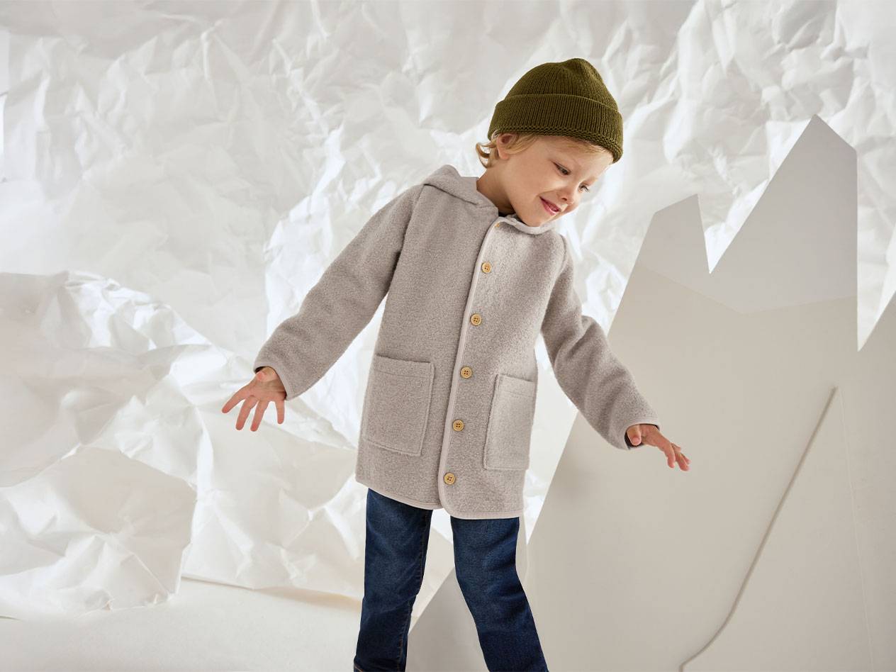 Dečak u sivoj vunenoj jakni i zelenoj kapi, nasmejan na pozadini zgužvanog papira.