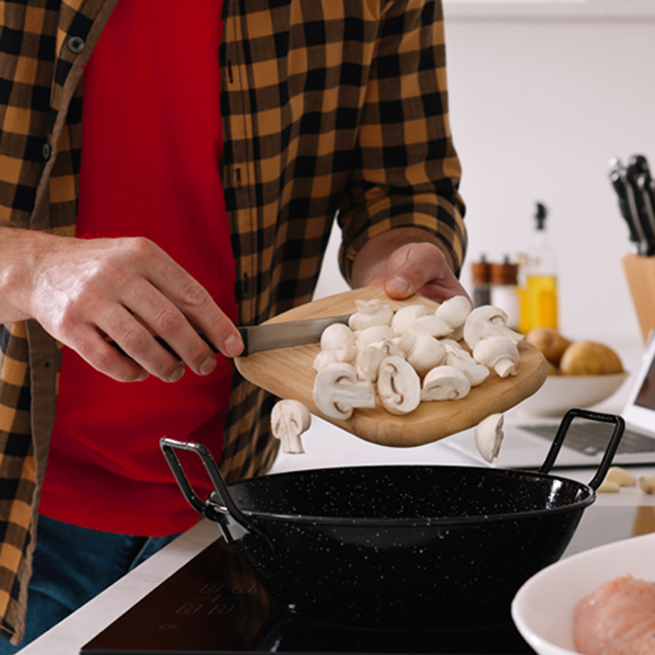 Muškarac sipa isečene pečurke sa daske u tiganj na šporetu.