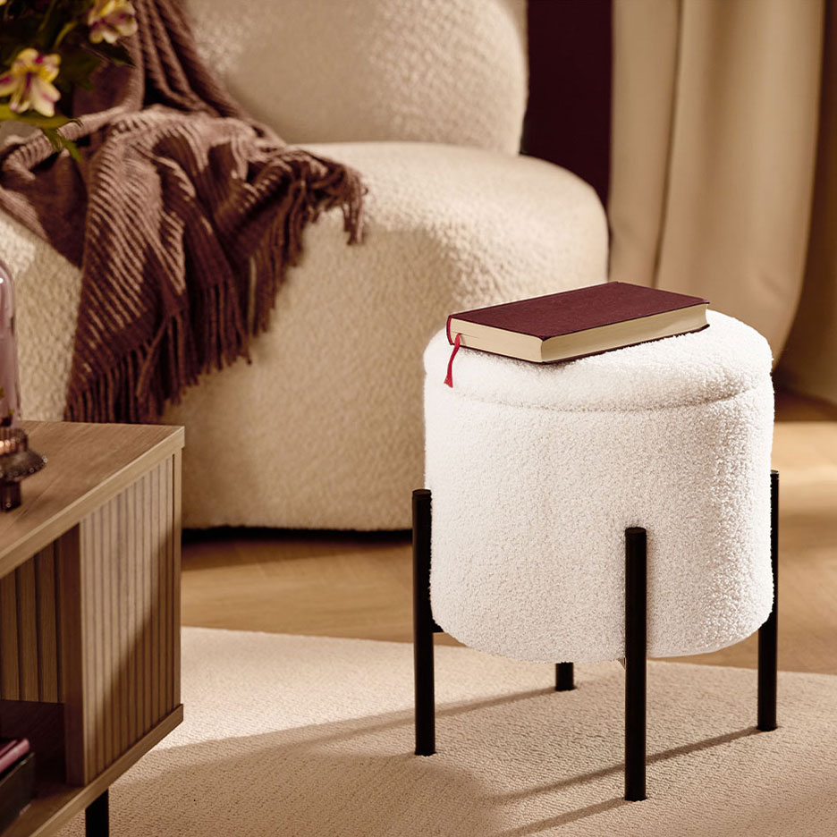 Bela pufnasta taburea sa crnim nogama, crvena knjiga, fotelja i braon ćebe.