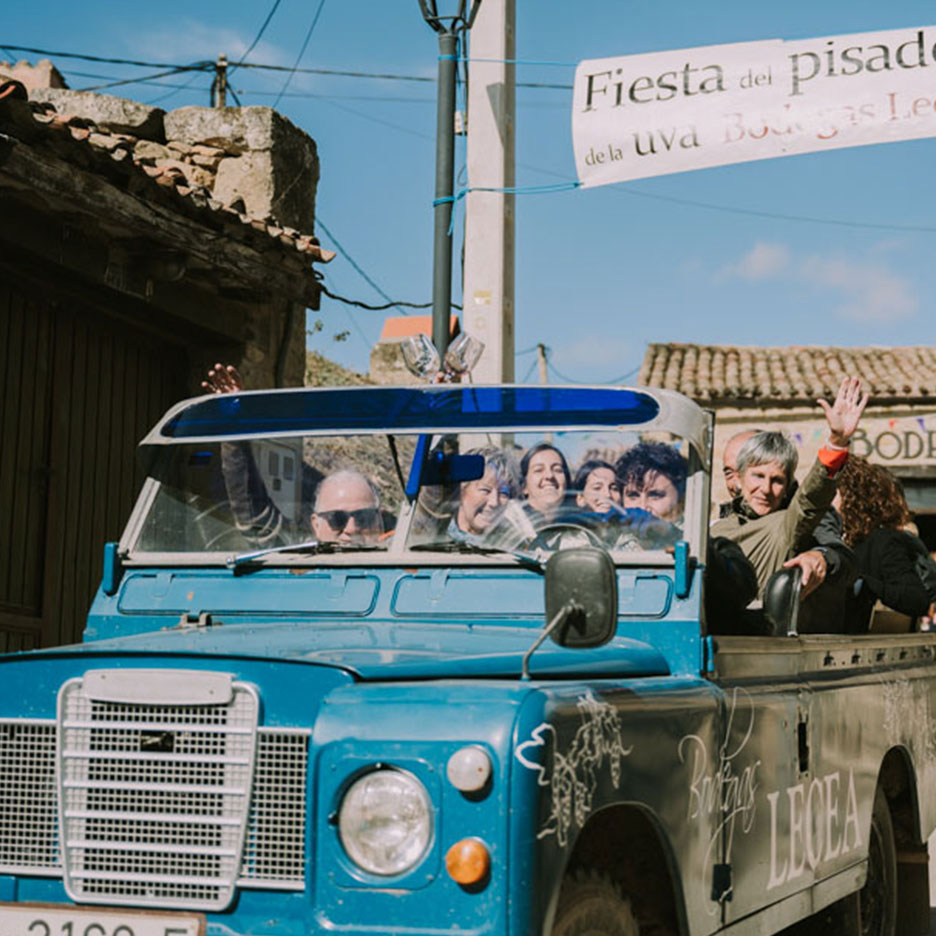 Ljudi mašu iz plavog terenskog vozila tokom vinskog festivala, sa transparentom „Fiesta del pisado de la uva“.