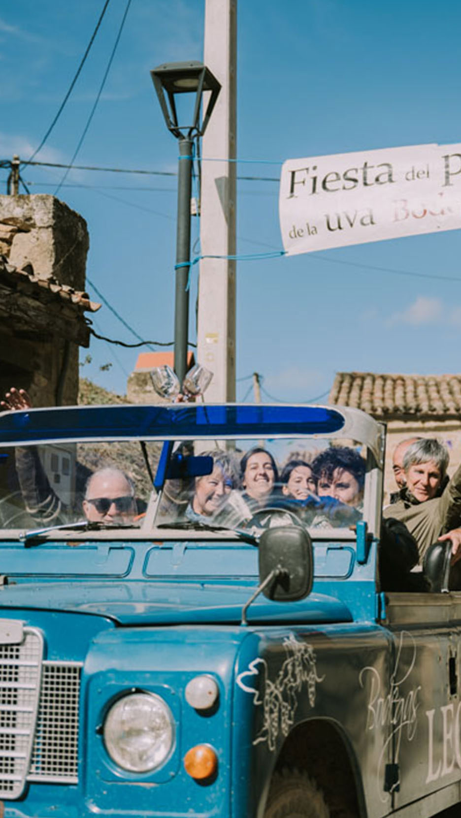 Ljudi u plavom automobilu ispod transparenta sa natpisom 'Fiesta del P de la uva Bod'.