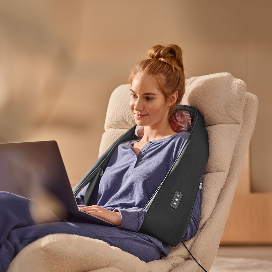 Žena sa masažerom za vrat i ramena radi na laptopu u udobnoj fotelji.