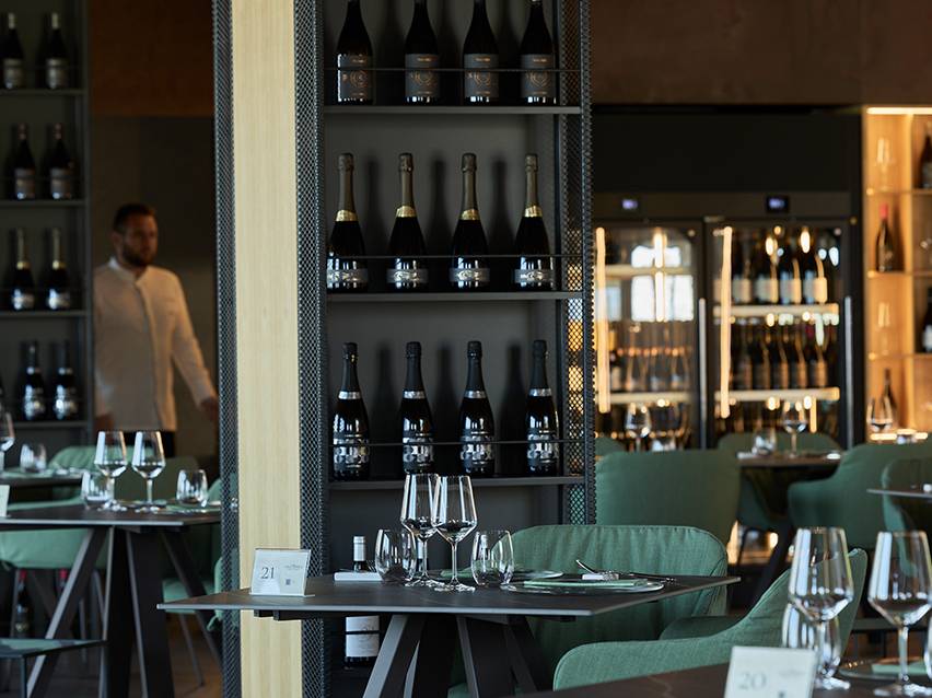 Enterijer restorana sa flašama vina na policama i stolovima postavljenim čašama.