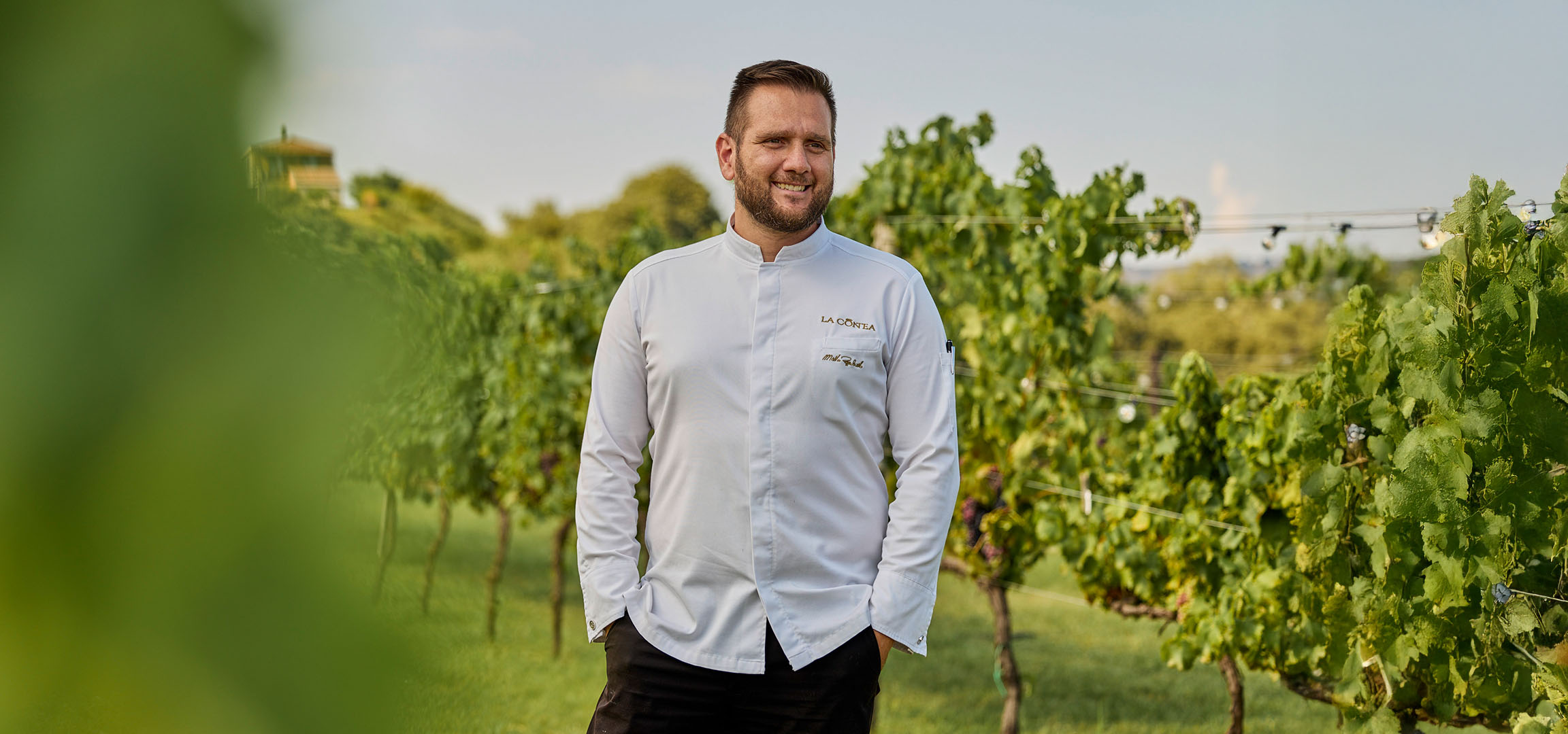 Nasmejani muškarac u beloj kuvarskoj jakni u sunčanom vinogradu.