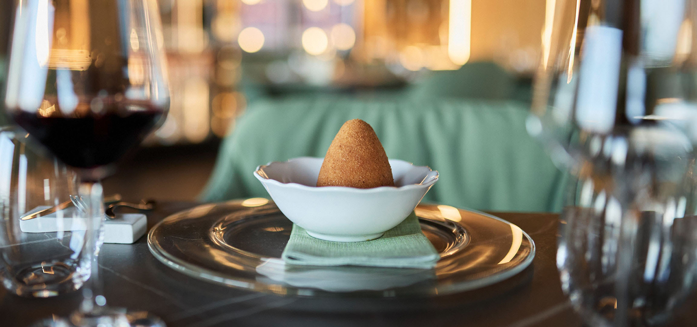 Zlatno konusno jelo u beloj činiji, sa crvenim vinom i priborom za jelo na stolu.