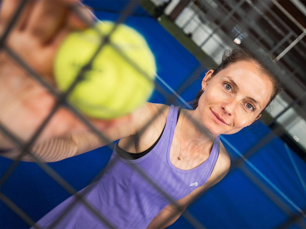 Žena u sportskoj odeći drži padel lopticu kroz mrežu, plavi teren u pozadini.