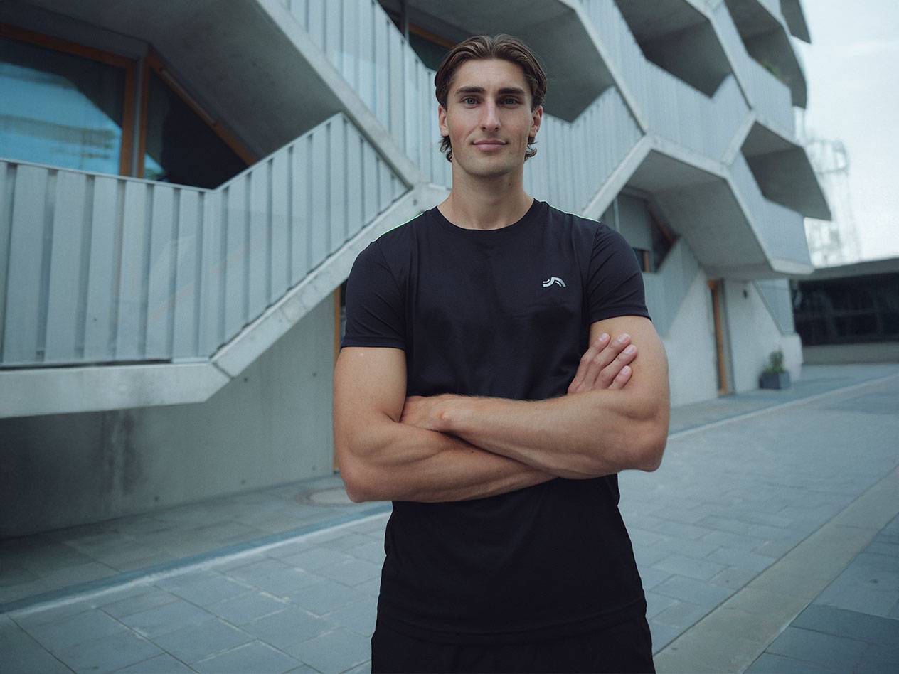 Muškarac u crnoj majici sa logotipom i crnim šortsevima, stoji napolju prekrštenih ruku.