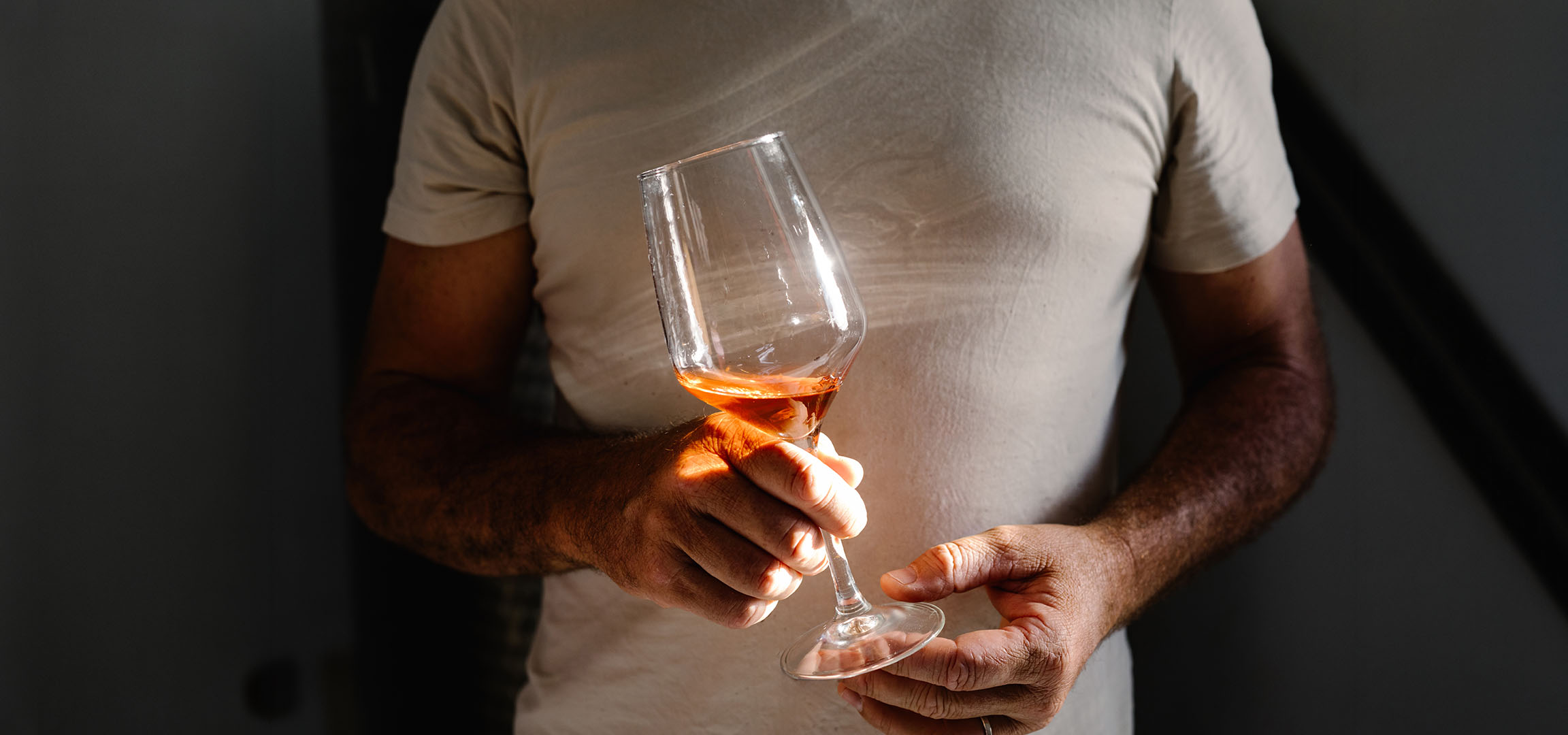 Muškarac drži čašu roze vina, obasjan suncem, na tamnoj pozadini.