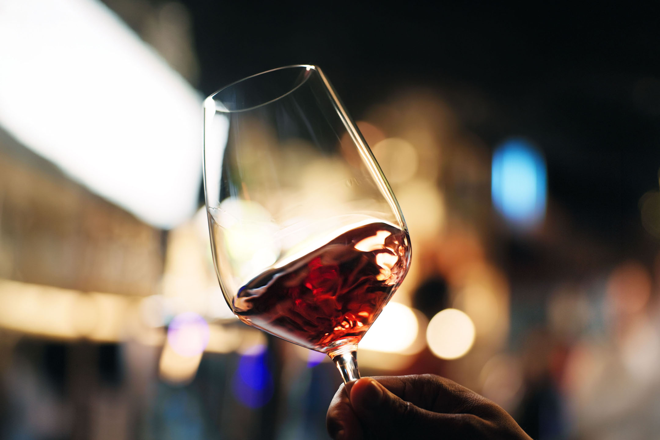 Čaša crvenog vina u ruci, sa zamagljenim svetlima u pozadini.