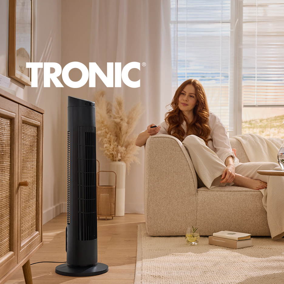 Žena se opušta na kauču sa daljinskim upravljačem, dok crni stubni ventilator stoji pored drvenog ormarića.