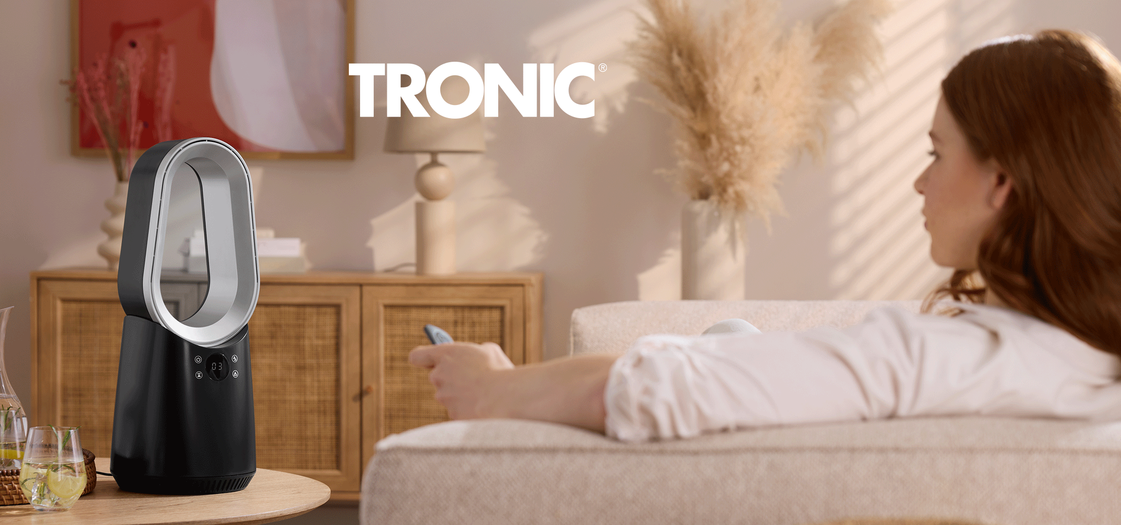 Žena koristi daljinski upravljač za crni ventilator bez lopatica, dok beli toranj ventilator stoji u dnevnoj sobi.