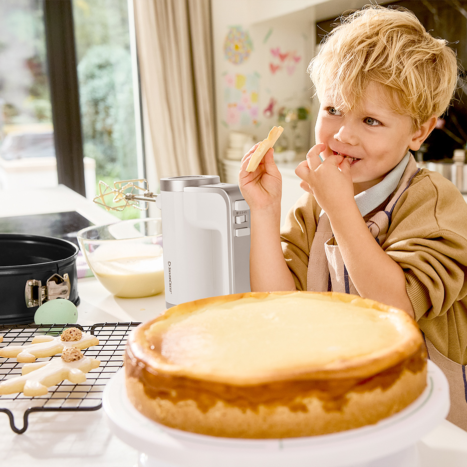Dečak jede kolačiće pored miksera i torte od sira u kuhinji.