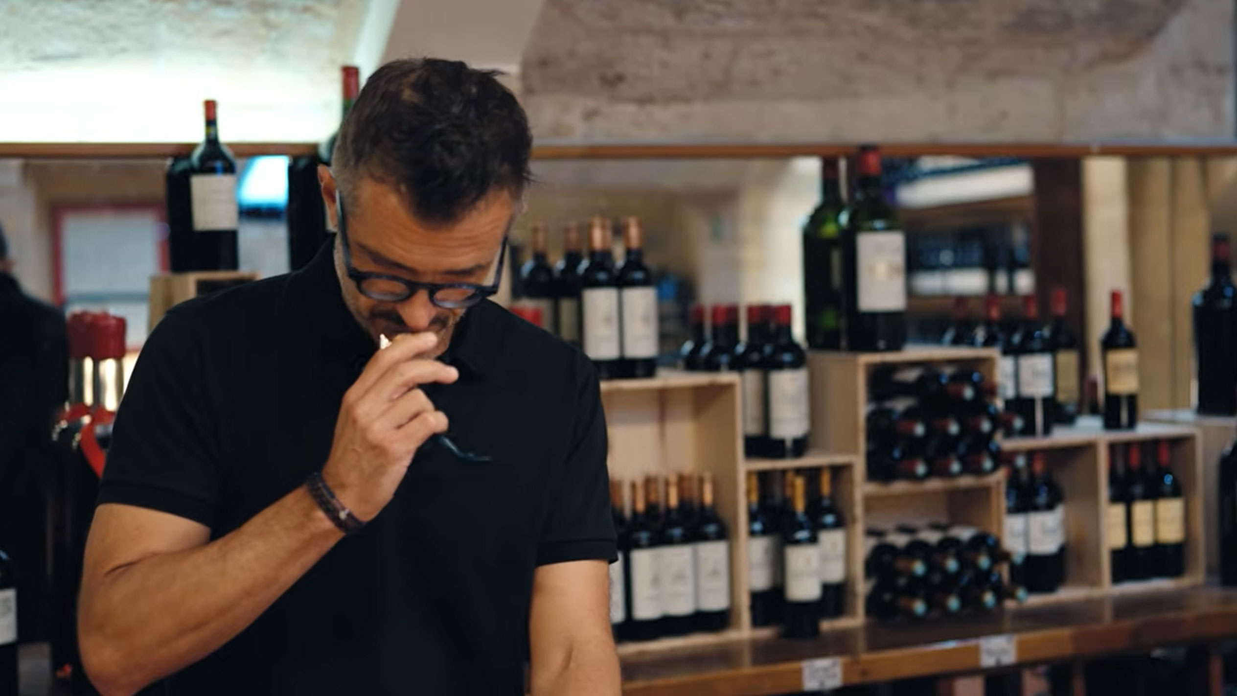 Muškarac u vinskom podrumu degustira vino, okružen flašama na drvenim policama.