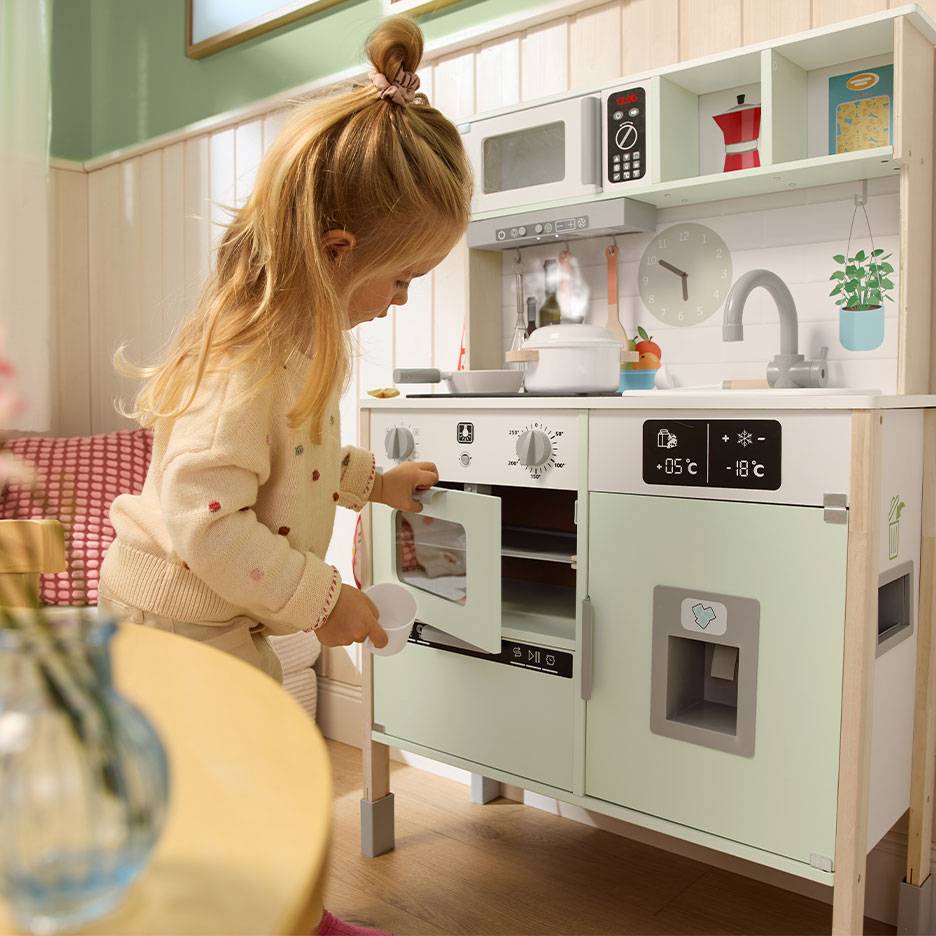 Dete se igra sa drvenom kuhinjom igračkom, sa rernom, mikrotalasnom i frižiderom.