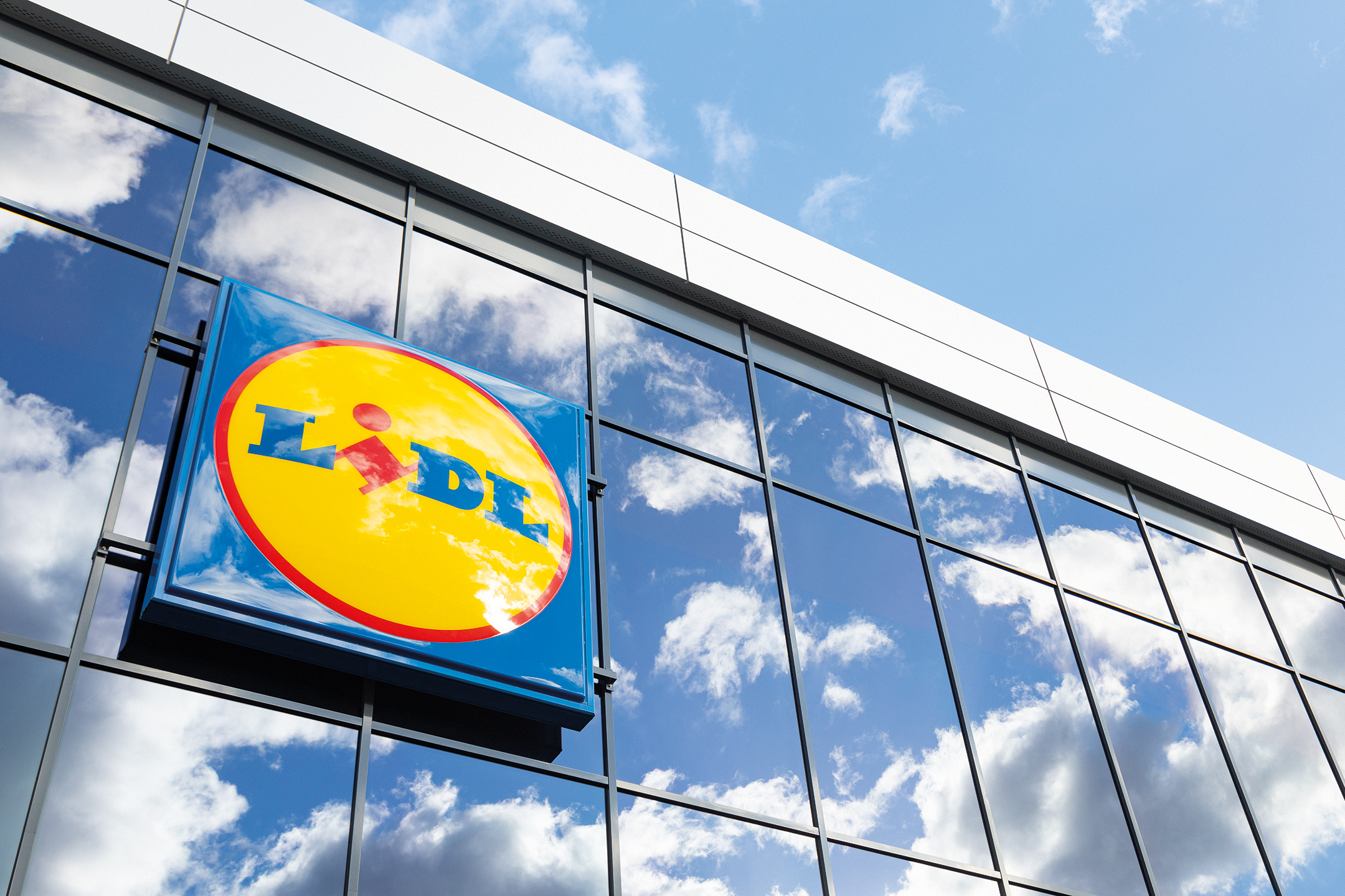 Lidl natpis na staklenoj fasadi zgrade, sa odrazom neba i oblaka.