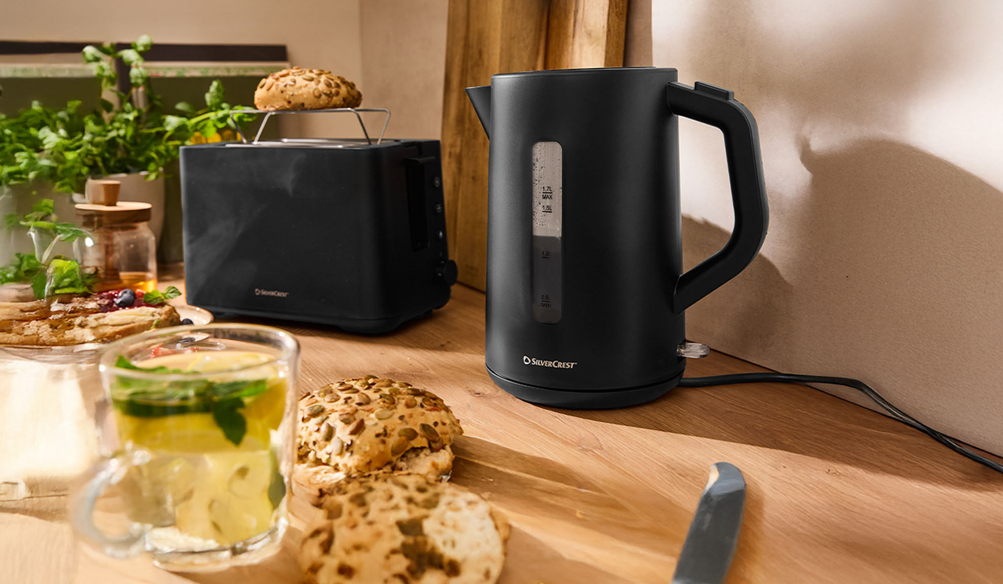 Crni SilverCrest čajnik i toster na drvenom stolu sa doručkom.