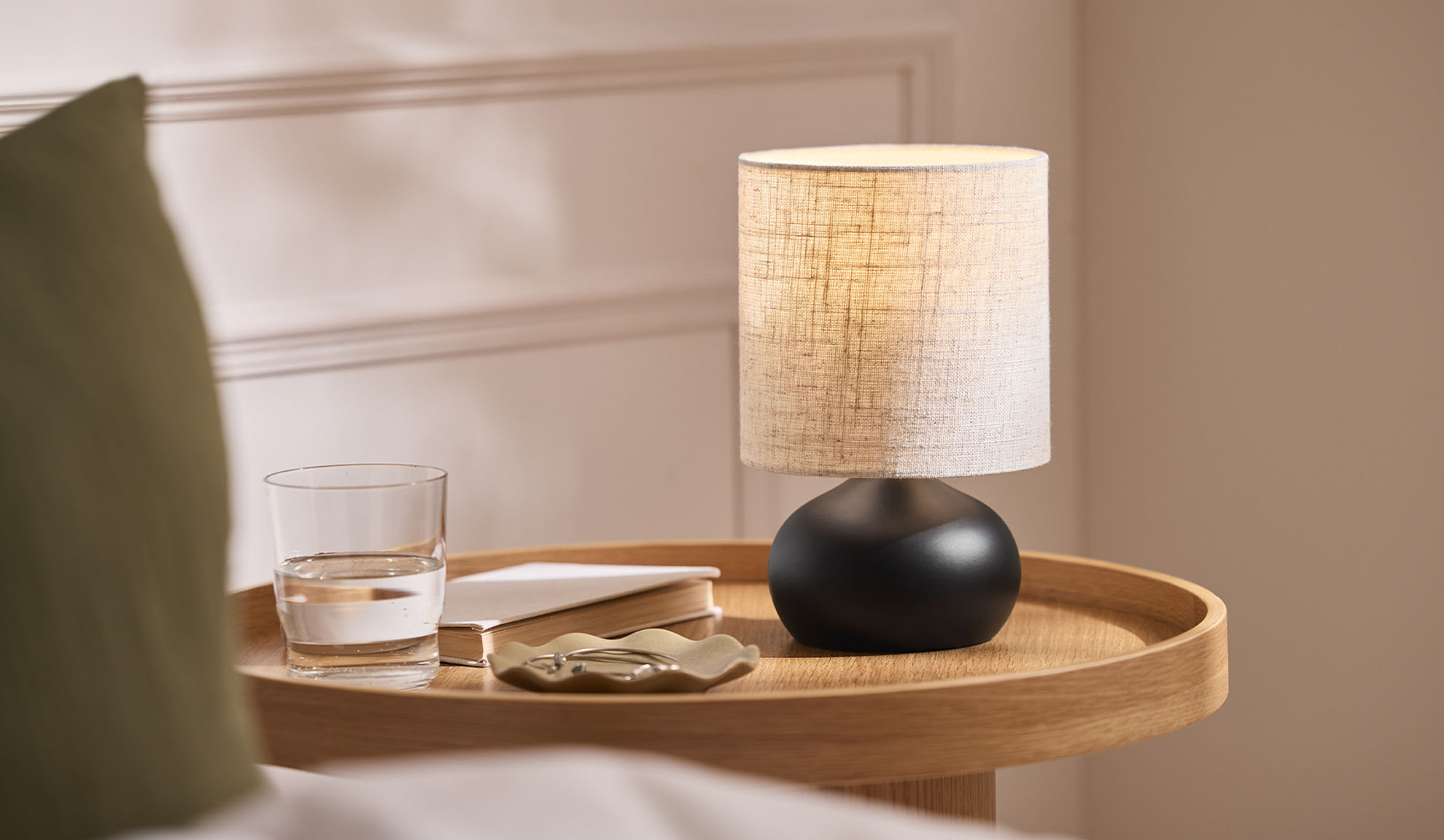 Noćna lampa, čaša, knjiga i poslužavnik za nakit na drvenom poslužavniku pored kreveta.