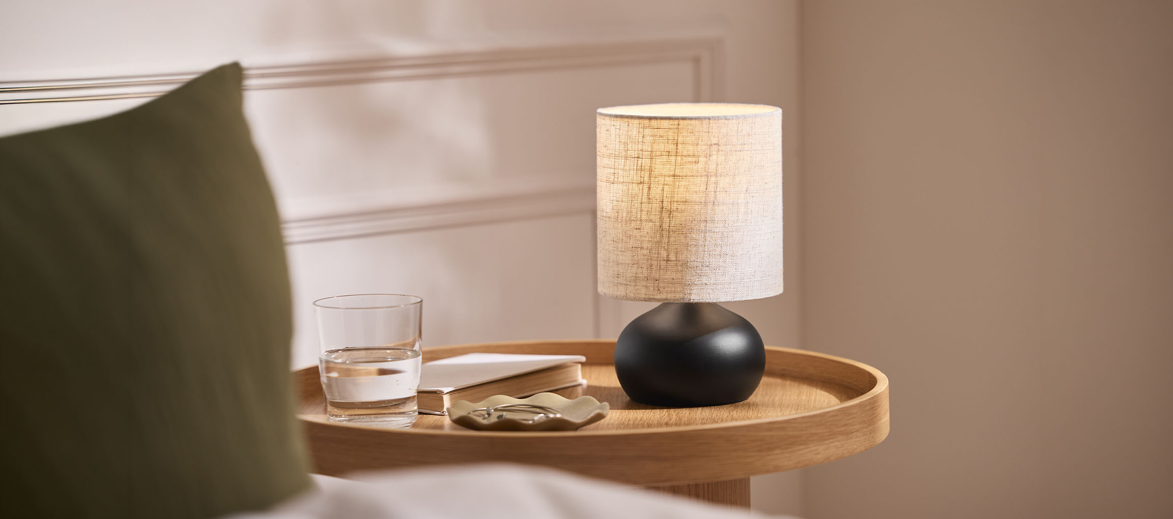 Noćna lampa sa platnenim abažurom, čaša vode i knjiga na drvenoj tacni.