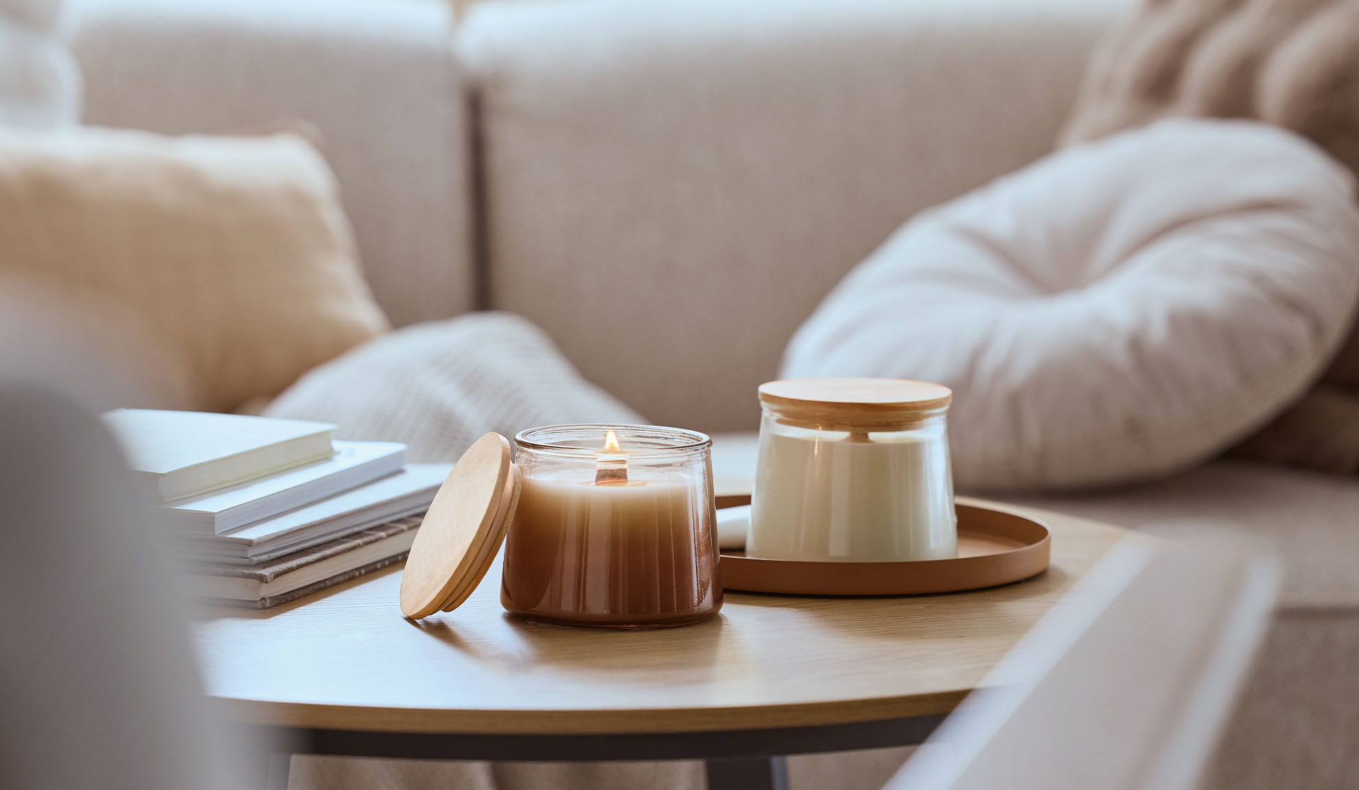 Dve mirišljave sveće na drvenom stolu, jedna upaljena, sa udobnom sofom u pozadini.