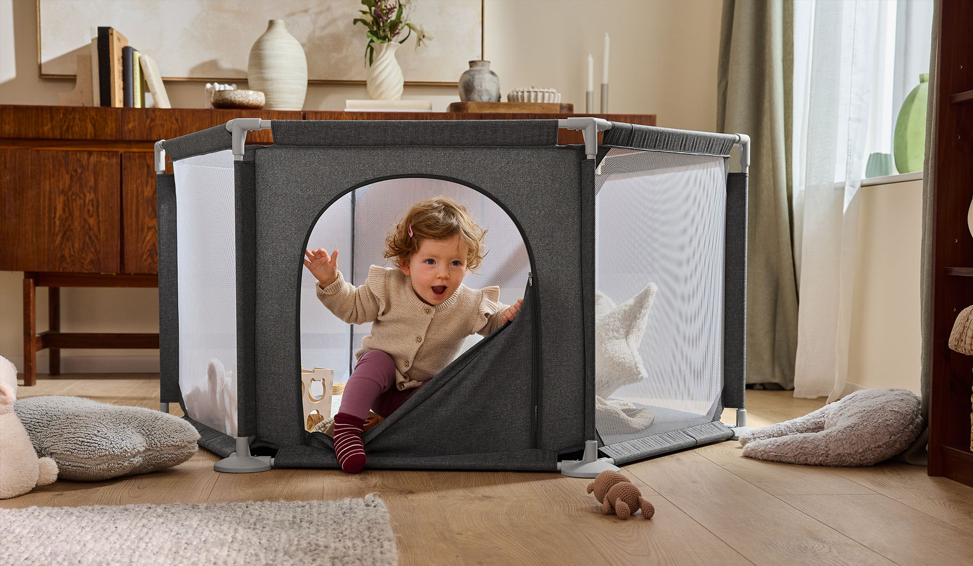 Srećna beba izlazi iz sivog ogradice u udobnoj dnevnoj sobi.