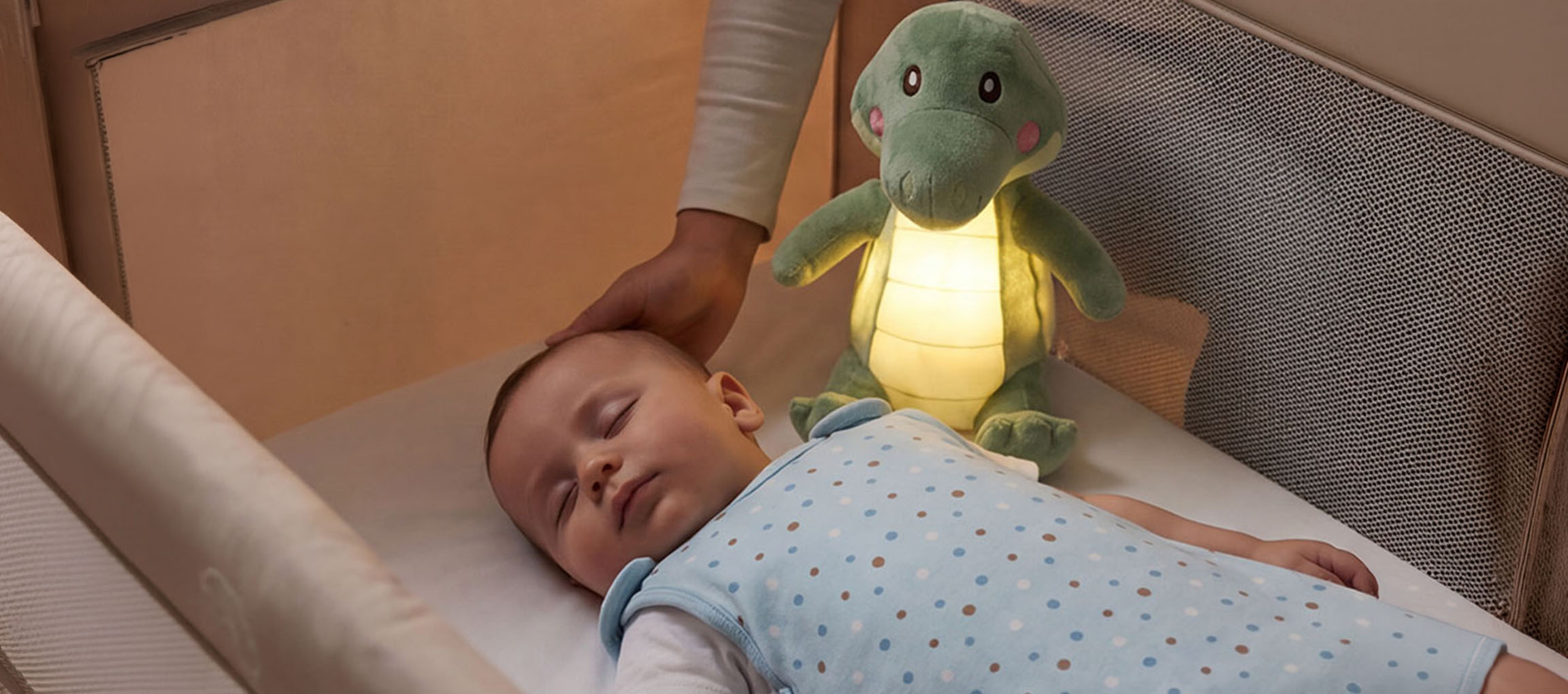 Beba spava u krevetiću sa plišanom noćnom lampom, odrasla osoba mazi bebinu glavu.